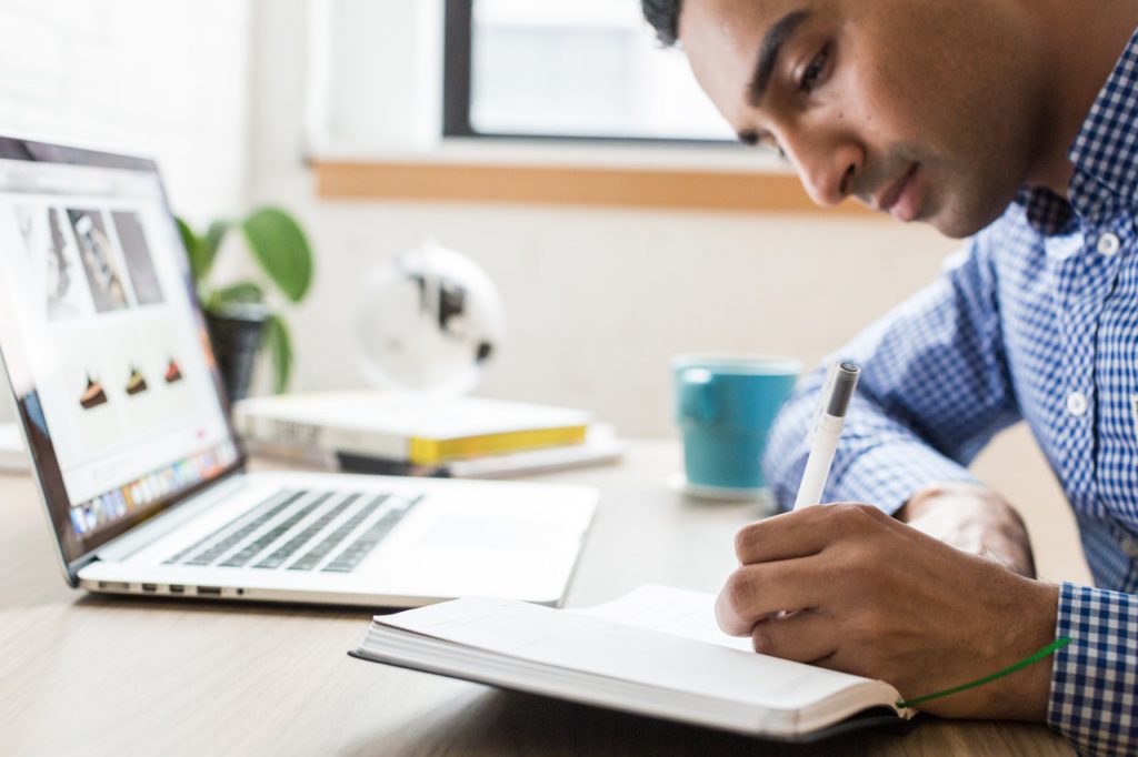 Empresário escrevendo em caderno dicas sobre gestão financeira com um notebook em sua frente