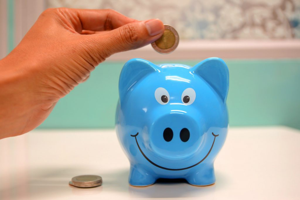 Um cofre azul no formato de um porquinho sobre uma mesa e uma mão feminina insere moedas