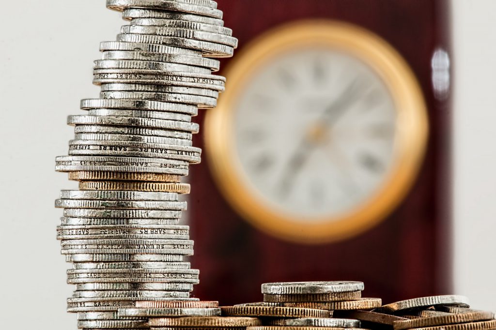 Relógio na parede desfocado com um molho de moedas à sua frente indicando a necessidade de fazer gestão financeira
