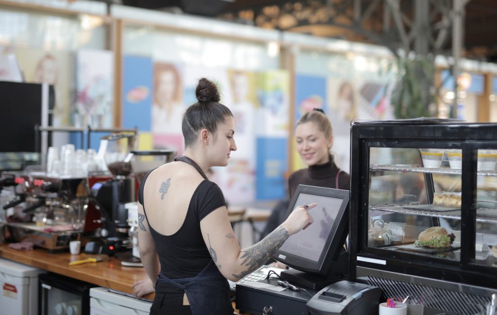 Mulher com tatuagens realizando atendimento à uma cliente em uma cafeteria