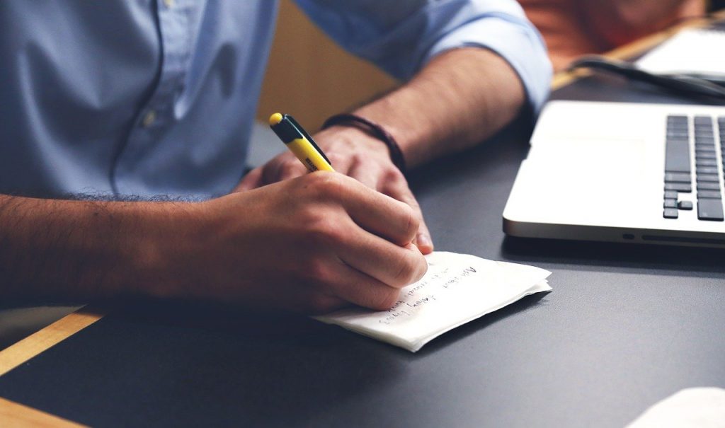 Homem escrevendo em um pedaço de papel em frente à um notebook, ele está iniciando seu planejamento financeiro